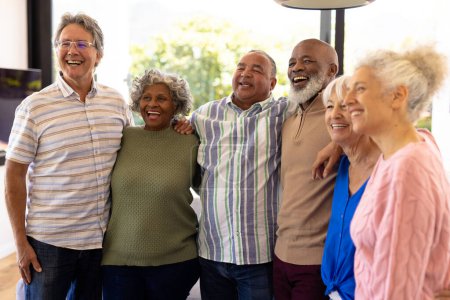 Multiracial senior friends with arms around laughing and looking away while standing in nursing home. Cheerful, unaltered, friendship, togetherness, support, assisted living and retirement concept.