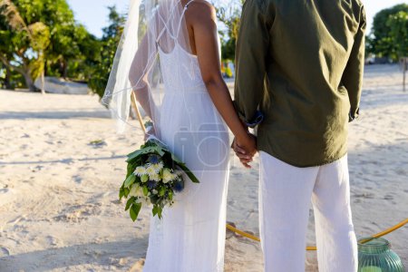 Midsection of caucasian newlywed couple holding hands and standing at beach at wedding ceremony. Unaltered, love, together, destination wedding, event, tradition, celebration, bouquet, hands, nature.