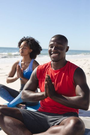 Glückliches, fittes afrikanisch-amerikanisches Paar, das am sonnigen Strand Yoga-Meditation praktiziert. Sommer, gesunder Lebensstil, Entspannung, Wohlbefinden und Urlaub, unverändert.