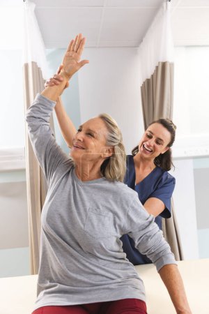 Caucasian female physiotherapist and senior woman stretching arm at hospital. Hospital, physiotherapy, work, medicine and healthcare, unaltered.