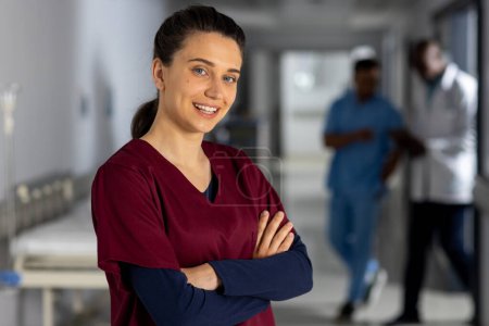 Portrait of happy caucasian female doctor wearing scrubs in corridor at hospital. Hospital, medicine, healthcare and work, unaltered.