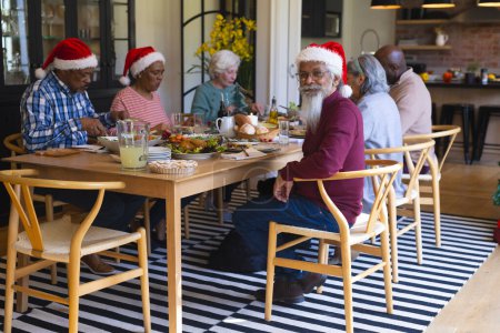 Happy diverse group of senior friends celebrating at christmas dinner in sunny dining room. Retirement, friendship, christmas, celebration, meal, senior lifestyle, communication unaltered.
