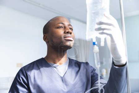African american male doctor wearing scrubs applying drip in hospital room. Medicine, healthcare, work and hospital, unaltered.