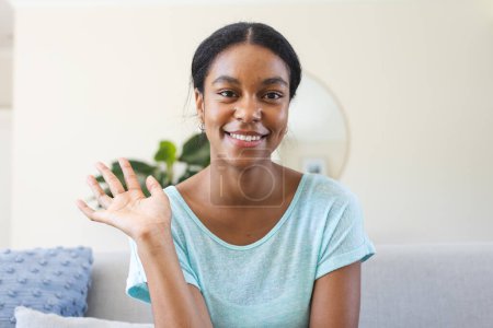 Young biracial woman waves cheerfully on video call, seated indoors. Her bright smile adds warmth to the cozy home setting.