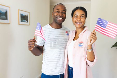 Diverse couple holding American flags at home, proudly wearing 'I Voted' stickers on their vote badges. They show civic pride, encouraging democratic participation.