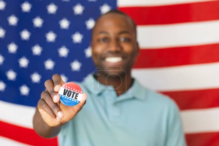 Young African American man shows a 'Vote' button. He stands before an American flag, symbolizing civic engagement and patriotism.