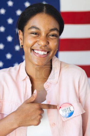 Young biracial woman smiles proudly in front of an American flag, showcasing her civic engagement with a "Vote" badge on her shirt.