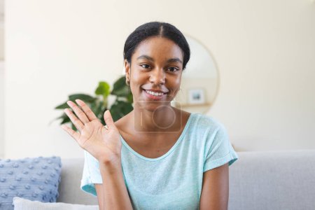 Young biracial woman waves cheerfully from her home on a video call. She exudes a friendly demeanor, comfortably seated in a cozy living room setting.