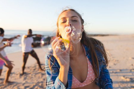 Young Caucasian woman blows bubbles on a sunny beach. Friends enjoy a playful moment in the background, highlighting leisure at the seaside.