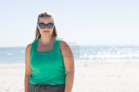Die junge Plus-Size-Kaukasierin steht selbstbewusst an einem sonnigen Strand mit Kopierraum. Ihr lässiges Outfit suggeriert einen entspannten Tag am Meer, unverändert.