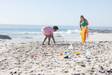 La joven mujer caucásica y el hombre afroamericano limpian una playa recogiendo basura con espacio para copiar. Demuestran responsabilidad ambiental recogiendo basura al aire libre.