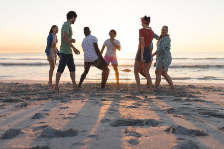 Vielfältige Freunde genießen einen Strandausflug bei Sonnenuntergang. Lachen und Gespräche erfüllen die Luft, während sie das Küstenambiente genießen.