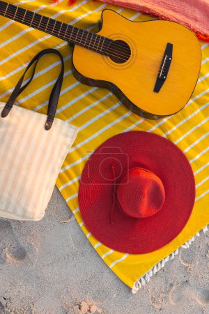 A beach setup features a guitar and a red hat on a yellow blanket. Beach essentials create a relaxing outdoor scene, evoking leisure and musical enjoyment.