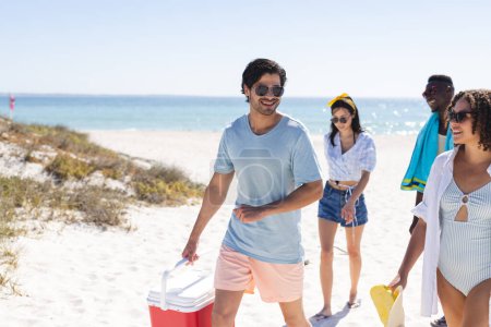 A diverse group of friends enjoys a sunny day at the beach. They carry beach gear, embodying a relaxed, outdoor leisure vibe.