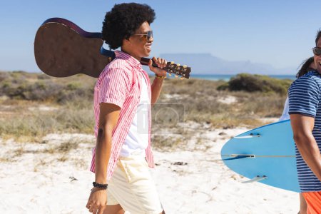 Un grupo diverso de jóvenes amigos disfruta de un día de playa soleado sin cambios. Un hombre afroamericano lleva una guitarra, acompañado de amigos de varias etnias, uno sosteniendo una tabla de surf, todo listo para la diversión al aire libre.