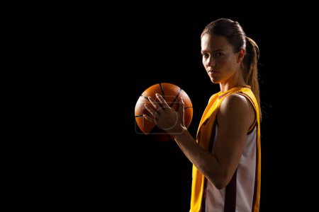 Young Caucasian female basketball player holds a basketball confidently on a black background, with copy space. She's in a dark setting, highlighting her focus and determination in the sport.