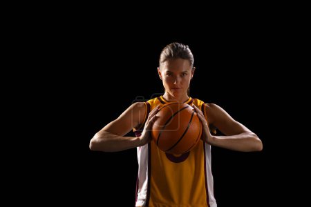 Junge kaukasische Basketballspielerin hält selbstbewusst einen Basketball auf schwarzem Hintergrund. Sie ist in einem dunklen Hintergrund, was ihren Fokus und ihre Entschlossenheit im Sport unterstreicht.