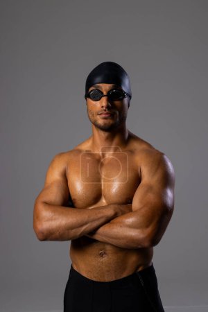 Athletic biracial male swimmer poses confidently in swimwear. His swim cap and goggles suggest he's a professional ready for competition.