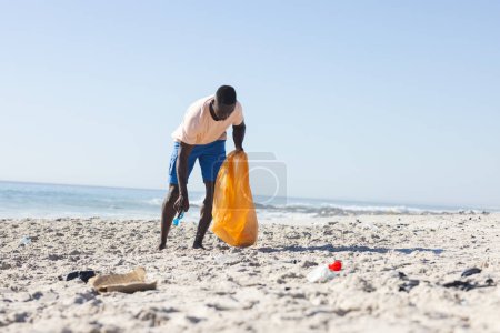 A young African American man cleans a beach, collecting trash, with copy space. He is actively participating in an environmental conservation effort outdoors.