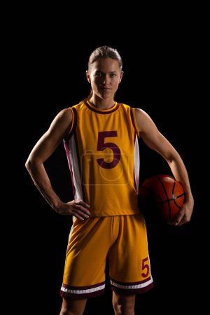 Confident female basketball player poses with a ball on a black background. Her athletic attire suggests she's ready for a competitive game on the court.