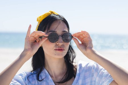 Eine junge Frau passt ihre Sonnenbrille am Strand unverändert an. Ihr entspanntes Auftreten lässt auf einen entspannten Tag im Freien schließen.
