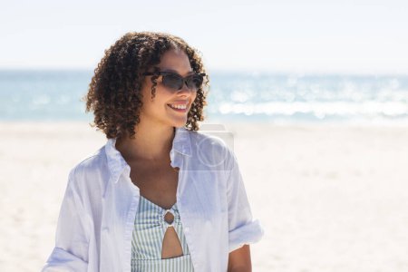 Young biracial woman enjoys a sunny beach day. She exudes happiness in a casual outfit, unaltered, with the ocean as a serene backdrop.