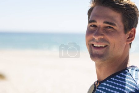 Junger Kaukasier lächelt am Strand, mit Kopierraum. Sein entspannter Gesichtsausdruck suggeriert einen entspannten Tag im Freien am Meer, unverändert.