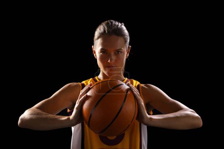 Young Caucasian female basketball player poses confidently in basketball attire on a black background. She exudes determination and strength, ready for a competitive game.