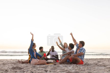 Diverse group of friends enjoy a beach sunset and having a party, with copy space. Laughter and high-fives create a relaxed outdoor atmosphere.