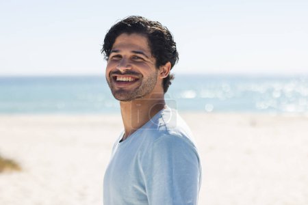 A young biracial man smiles brightly at the beach. His casual attire and joyful expression suggest a relaxed outdoor setting unaltered.