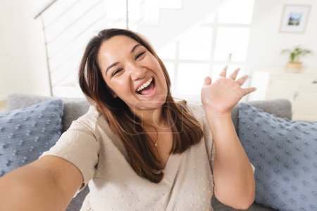 Young biracial woman greets warmly from her cozy home on a video call. Her cheerful expression adds a welcoming vibe to the living room setting.