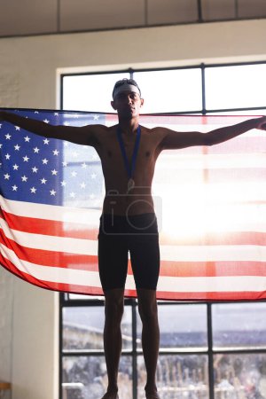 Stolze junge zweirassige männliche Schwimmer zeigen die amerikanische Flagge, mit Kopierraum. Er feiert einen sportlichen Sieg mit einer Medaille, die Leistung und Patriotismus symbolisiert.
