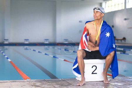 Swimmer wrapped in an Australian flag sits poolside with a medal, with copy space. The setting suggests a competitive atmosphere at an indoor swimming facility.