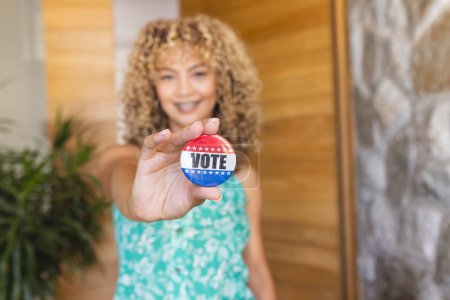Young biracial woman holding a 'VOTE' badge, with copy space. Her gesture promotes the importance of participating in elections.