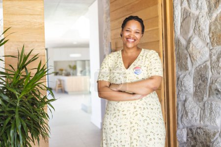 Young biracial woman stands confidently in a bright home setting, wearing a vote badge. Her nurse uniform suggests she's ready for a healthcare shift, radiating professionalism and care.