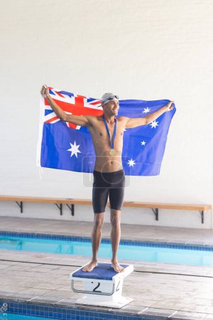Young biracial male swimmer celebrates victory at a swimming pool with the Australian flag. Holding the Australian flag, the athlete stands proud with a medal, symbolizing triumph.