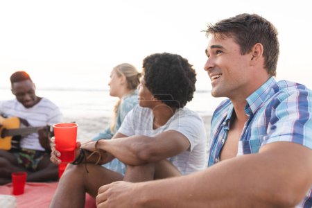 Vielfältige Freunde treffen sich am Strand. Sie teilen Lachen und Musik bei einem Picknick am Meer.