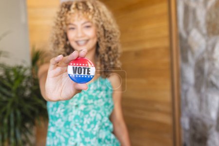 Young biracial woman holds a 'vote' badge towards the camera, with copy space. Her cheerful expression promotes the importance of participating in elections.