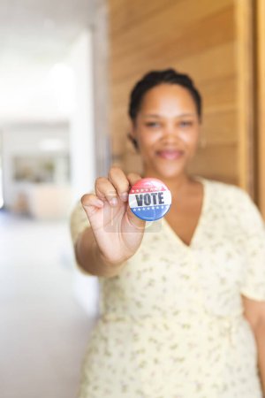 Young biracial woman holds a 'vote' badge, with copy space. Encouraging civic participation, she stands in a well-lit indoor setting.