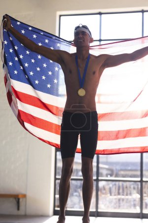 Young biracial male athlete swimmer celebrates victory with an American flag. Pride and accomplishment radiate from the swimmer's triumphant pose indoors.