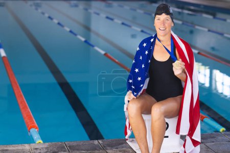 Caucasian woman celebrates at the poolside with an American flag, with medal, with copy space. Wrapped in the American flag, the athlete holds a gold medal, symbolizing victory.