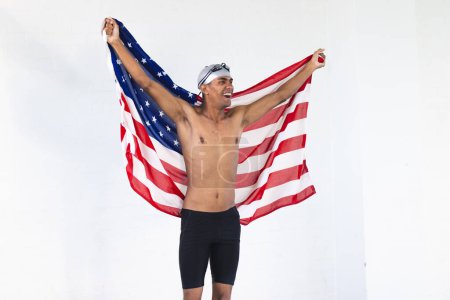 Junge zweirassige männliche Athleten Schwimmer feiert mit einer amerikanischen Flagge, mit Kopierraum. Sein Ausdruck von Freude und Patriotismus wird in einem hellen Rahmen deutlich.