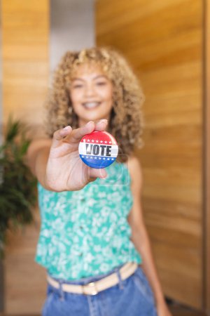 Young biracial woman shows a 'vote' button, with copy space. She encourages civic engagement and the importance of voting from home.