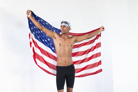 Young biracial male athlete swimmer holds an American flag with pride. His athletic build suggests a sports victory or patriotic celebration.
