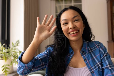 Young biracial woman waves at the camera from home on video call. She exudes a friendly vibe in a casual setting, creating a welcoming atmosphere.