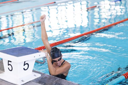 A swimmer celebrates victory at the poolside, with copy space. The athlete's raised arm and joyful expression convey a sense of triumph after a competitive race.
