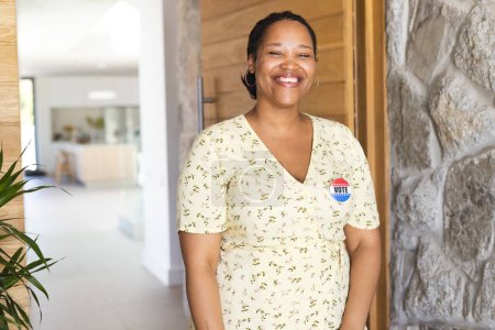 A smiling young biracial woman stands in a bright hallway, wearing a vote badge, with copy space. Her badge promotes civic engagement in a healthcare setting.