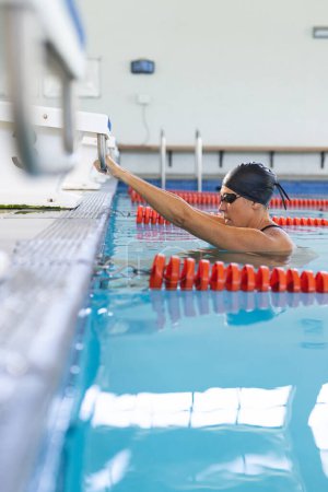 Kaukasische Athletenschwimmerin legt am Beckenrand eine Pause ein. Ihre Badekappe und ihre Brille deuten auf eine Trainingseinheit in einer Schwimmhalle hin..