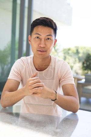 Young Asian man sits at a table on a video call. His casual attire suggests a relaxed setting at home or a casual office.