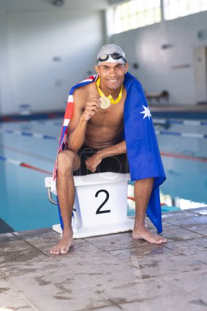 A young biracial male swimmer celebrates at the poolside, draped in an Australian flag with a medal. Pride shines on his face as he displays a hard-earned medal, symbolizing his achievement.